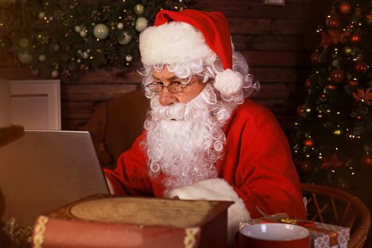 Santa Claus using laptop computer sitting in cozy room near fireplace