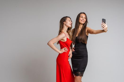 Happy beautiful two female friends posing in studio while making selfie on smartphone, isolated on grey background. Party concept