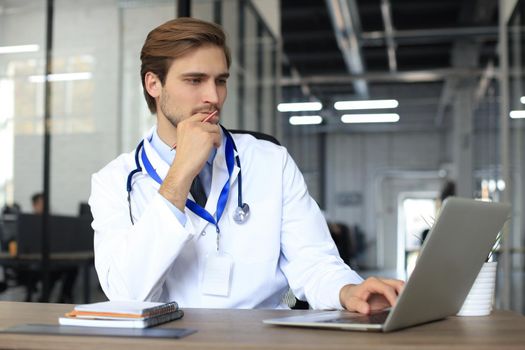 Doctor in white medical uniform with statoscope reading personal file of patient on tablet and writes data to laptop