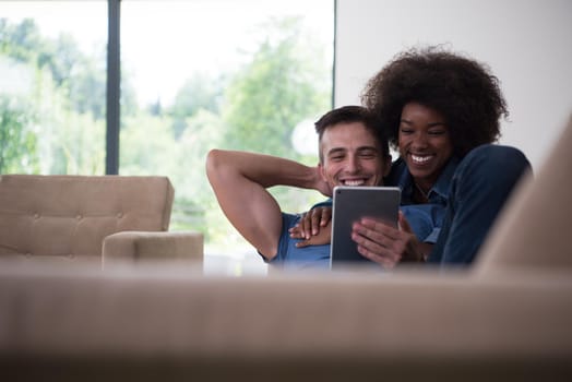 Young multiethnic couple relaxing at luxurious home with tablet computers reading in the living room on the sofa couch.