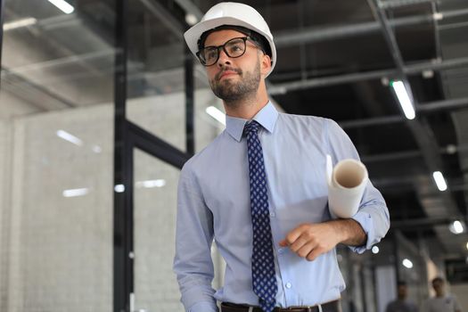 Confident young business man in hardhat holding blueprint and looking away while standing indoors
