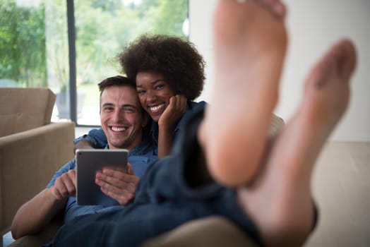 Young multiethnic couple relaxing at luxurious home with tablet computers reading in the living room on the sofa couch.