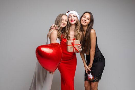 Pretty young girls friends in festive dresses holding gift, red balloon, champagne celebrating holiday on gray backdrop. High quality photo