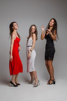 Charming positive young girls friends wearing stylish feminine evening dresses posing on gray studio background. High quality photo