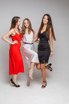Charming positive young girls friends wearing stylish feminine evening dresses posing on gray studio background. High quality photo