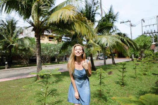 Young smiling woman wearing jeans sundress and standing near palms in background. Conept of resting in Florida and summer exotic vacations.