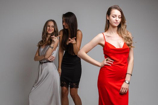 Studio portrait of gossiping girls in silver and black dresses mocking beautiful unhappy girl with long fair hair in red dress standing in the foreground and looking down sadly.