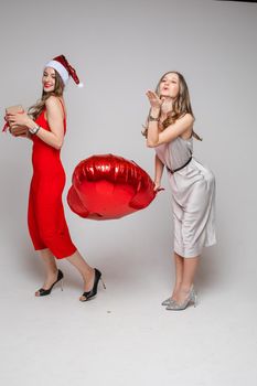 Happy two pretty women with heart-shaped balloon and gift box posing in studio, isolated on grey background. Party concept