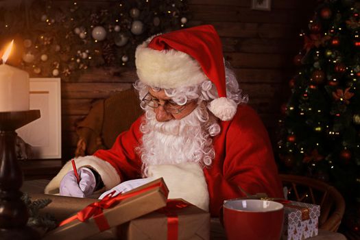 Portrait of happy Santa Claus sitting at his room at home near Christmas tree and answering Christmas letters