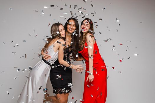 Celebrating young girls friends wearing evening dresses hugging under silver confetti on holiday party on gray backdrop. High quality photo