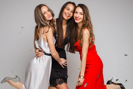 Portrait of three happy attractive girls in white, black and red silk dresses and heels hugging together and smiling at camera. Confetti falling down.
