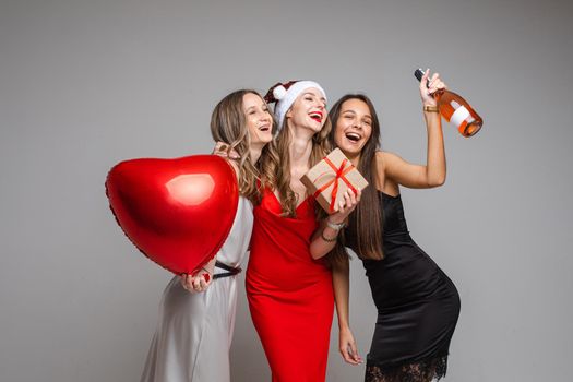 Studio portrait of gorgeous happy ladies in silk dresses with red heart air balloon, gift and bottle of rose wine having fun embracing and posing on grey background.
