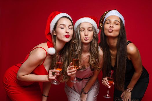 Portrait of three beautiful ladies in Santa hats and cocktail dresses holding glasses of wine and making pouting lips to the camera with eyes closed. Isolate on red.