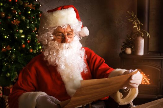 Portrait of happy Santa Claus sitting at his room at home near Christmas tree and reading Christmas letter or wish list