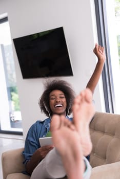 young beautiful African American woman enjoys listening to music with headphones and tablet in your armchair in her luxury home