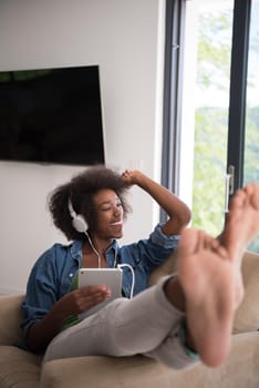 young beautiful African American woman enjoys listening to music with headphones and tablet in your armchair in her luxury home