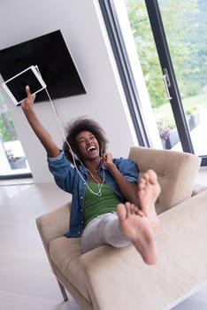 young beautiful African American woman enjoys listening to music with headphones and tablet in your armchair in her luxury home