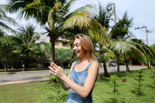 Youg caucasian woman talking by sartphone near palm trees and road, wearing jeans sundress. Concept of modern technology and summer vacations in tropical place, exotic atmospehere.