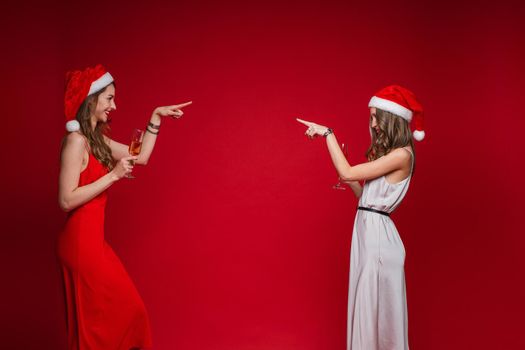 Side view of smiling pretty young women in santa hats pointing fingers at each other, isolated on red background. Party concept