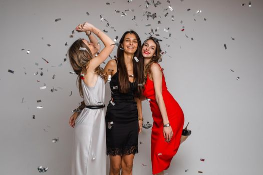 Celebrating young girls friends wearing evening dresses hugging under silver confetti on holiday party on gray backdrop. High quality photo