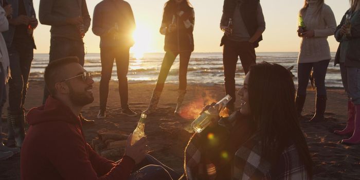 Happy Carefree Young Friends Having Fun And Drinking Beer By Bonefire On The Beach As The Sun Begins To Set