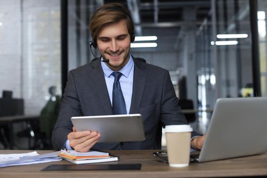 Cheerful young support phone male operator in headset, at workplace while using laptop, help service and client consulting call center concept.