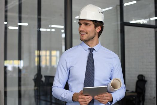 Confident young business man in hardhat holding blueprint and looking away while standing indoors