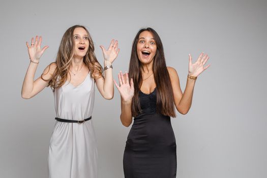 Portrait of two astonished girlfriends in silver and black dresses looking with wide eyes and half raised arms in shock and surprise. Isolate on white background.