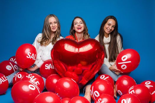 Happy women friends posing with red heart shaped balloon and air balls with percent and sale lettering on blue backdrop. High quality photo