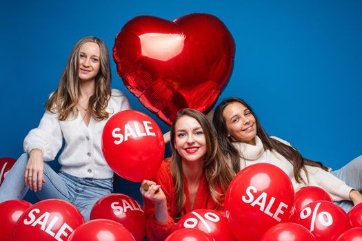 Happy pretty female friends sitting with sale red air balloons, isolated on blue background. Shopping holiday concept