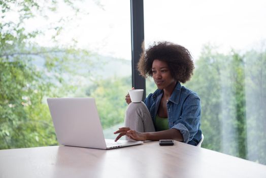 Young african american woman smiling sitting near bright window while looking at open laptop computer on table and holding white mug in her luxury home