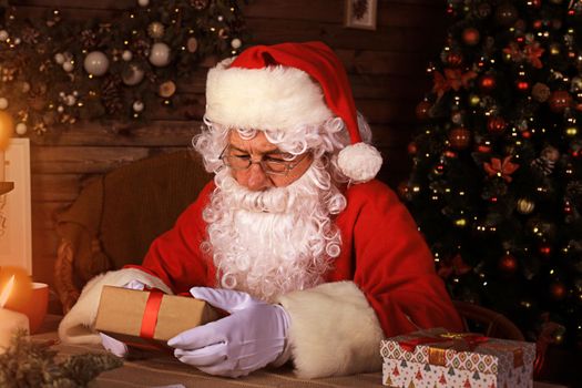 Portrait of happy Santa Claus sitting at his room at home near Christmas tree with gift box