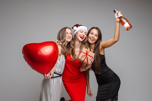 Studio portrait of gorgeous happy ladies in silk dresses with red heart air balloon, gift and bottle of rose wine having fun embracing and posing on grey background.