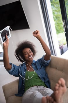 young beautiful African American woman enjoys listening to music with headphones and tablet in your armchair in her luxury home