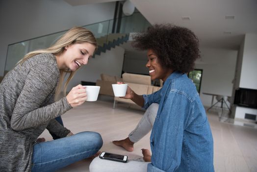 Two young laughing multiethnic women sit on the floor and enjoy while drinking coffee using smartphone in luxury home