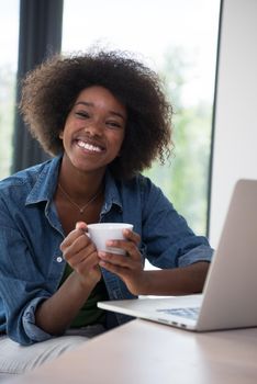Young african american woman smiling sitting near bright window while looking at open laptop computer on table and holding white mug in her luxury home