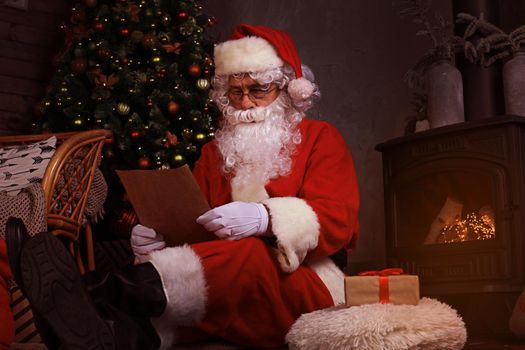 Portrait of happy Santa Claus sitting at his room at home near fireplace and reading Christmas letter or wish list