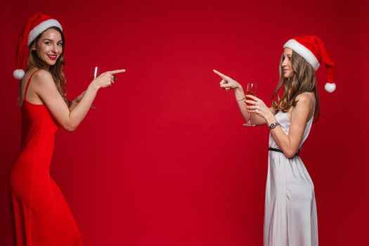 Side view of smiling pretty young women in santa hats pointing fingers at each other, isolated on red background. Party concept