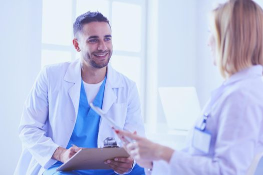 Handsome doctor is talking with young female doctor and making notes while sitting in his office