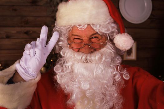 Traditional Santa Claus standing by the Christmas tree in a room and showing sign Victory