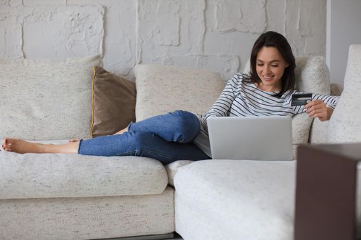 Happy natural brunette using credit card and laptop in the living room