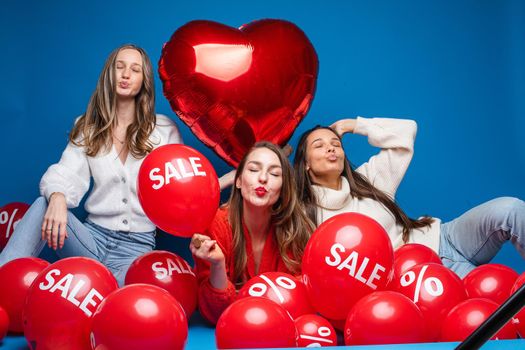 Portrait of three pretty smiling girlfriends in casual clothes sitting with red air balloons with sale and percent sign. Girl in the middle holding red heart-shaped balloon. Cutout on blue.