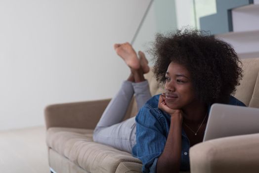 Smiling young African American woman using laptop while relaxing on sofa at luxury home