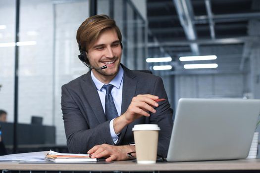 Smiling male call-center operator with headphones sitting at modern office, consulting online information in a laptop, looking up information in a file in order to be of assistance to the client