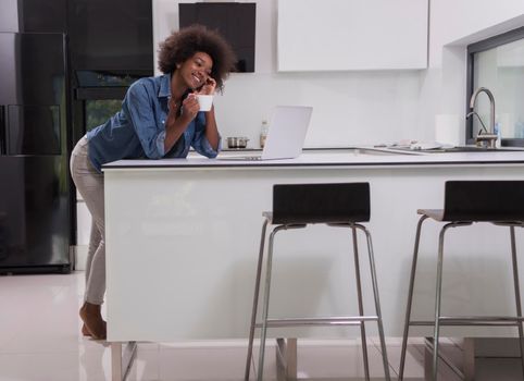 Young smiling black woman using computer and drinking coffee in modern kitchen interior