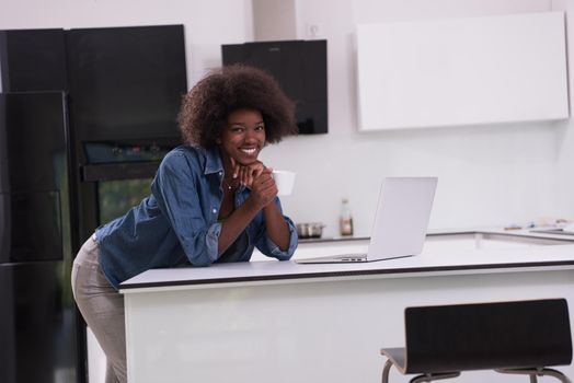Young smiling black woman using computer and drinking coffee in modern kitchen interior