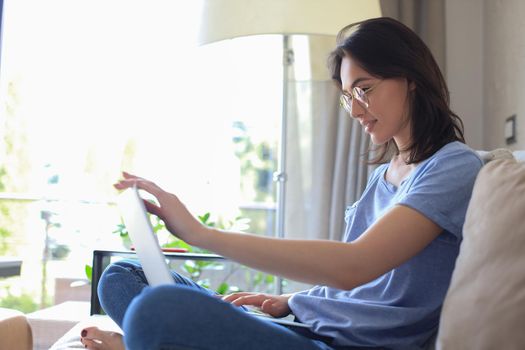 Smiling young woman sitting on sofa with laptop computer and chating with friends
