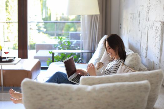 Smiling young woman sitting on sofa with laptop computer and chating with friends, little dog playing near