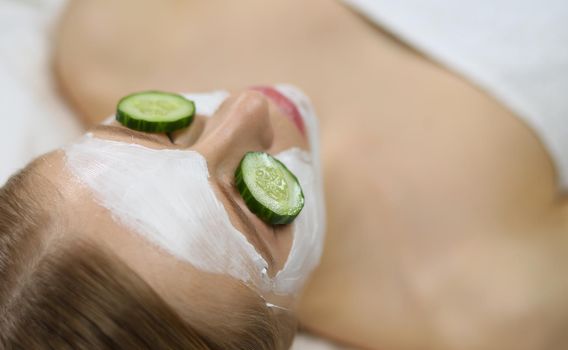 Beautiful young woman receiving facial mask of cucumber in beauty salon .