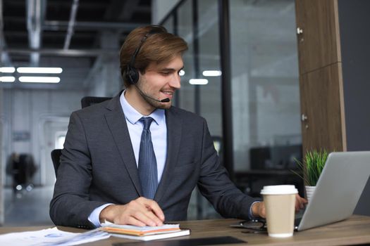 Smiling male call-center operator with headphones sitting at modern office, consulting online information in a laptop, looking up information in a file in order to be of assistance to the client